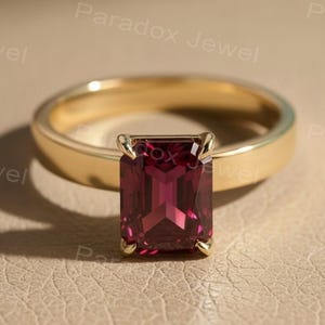 May include: A gold ring with a rectangular, deep red gemstone. The ring is set on a neutral-toned surface. The text "Paradox Jewel" is visible in the background.