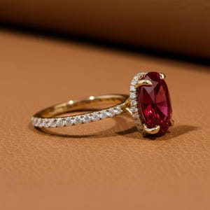 Może przedstawiać: Złoty pierścionek z dużym, owalnym rubinem. Obręcz pierścionka zdobią małe, przezroczyste kamienie szlachetne, a rubin otoczony jest aureolą mniejszych kamieni. Pierścionek jest prezentowany na brązowym tle.