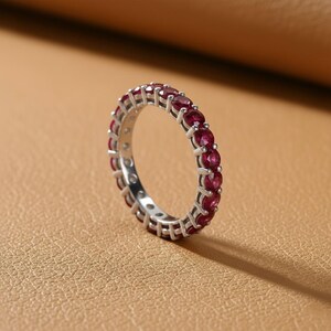 May include: A silver eternity ring with a continuous row of vibrant red gemstones. The ring is set against a neutral background, highlighting the sparkle of the gemstones. The ring's design is simple yet elegant, showcasing the beauty of the red stones.