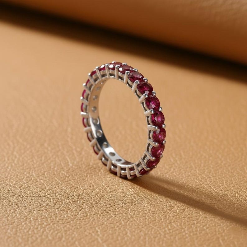 May include: A silver eternity ring with a continuous row of vibrant red gemstones. The ring is set against a neutral background, highlighting the sparkle of the gemstones. The ring's design is simple yet elegant, showcasing the beauty of the red stones.