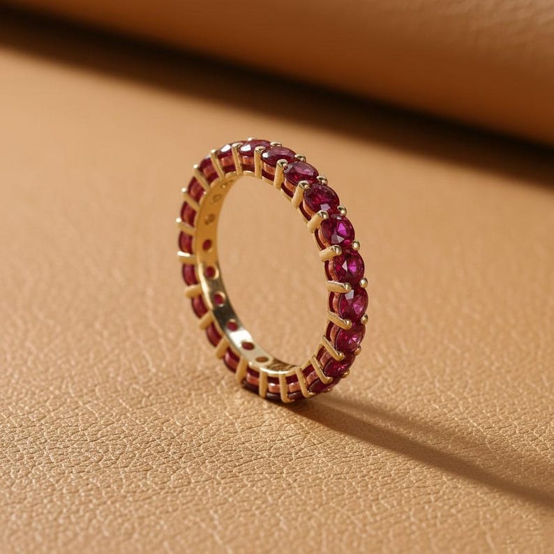May include: A gold-colored ring with a continuous band of small, round, red gemstones. The ring is set against a neutral, textured background, highlighting the vibrant color of the stones and the ring's design.