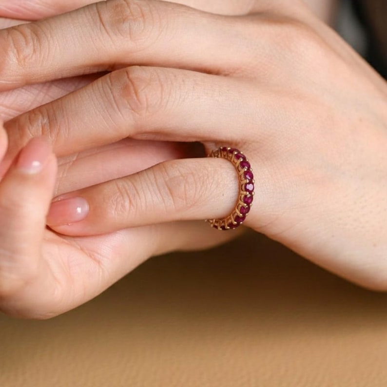 May include: A gold ring set with a continuous line of small, round, red gemstones. The ring is on a finger, with the hand and part of the arm in view. The ring's design is simple, with the gemstones closely set.