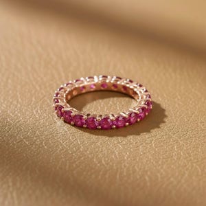 May include: A rose gold eternity ring with a continuous row of vibrant pink gemstones. The ring is set against a warm, neutral background, highlighting the sparkle of the gemstones. The ring's design is simple and elegant.