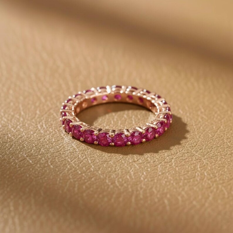 May include: A rose gold eternity ring with a continuous row of vibrant pink gemstones. The ring is set against a warm, neutral background, highlighting the sparkle of the gemstones. The ring's design is simple and elegant.