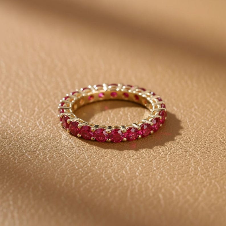 May include: A gold-colored eternity ring with a continuous row of vibrant red gemstones. The ring is set against a warm, neutral background, highlighting the sparkle of the gemstones. This piece of jewelry is a classic and elegant accessory.