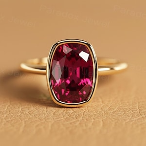 May include: A gold ring featuring a large, rectangular, deep red gemstone. The gemstone is set in a gold bezel, and the band is a simple, polished gold. The ring is displayed on a neutral, tan background.