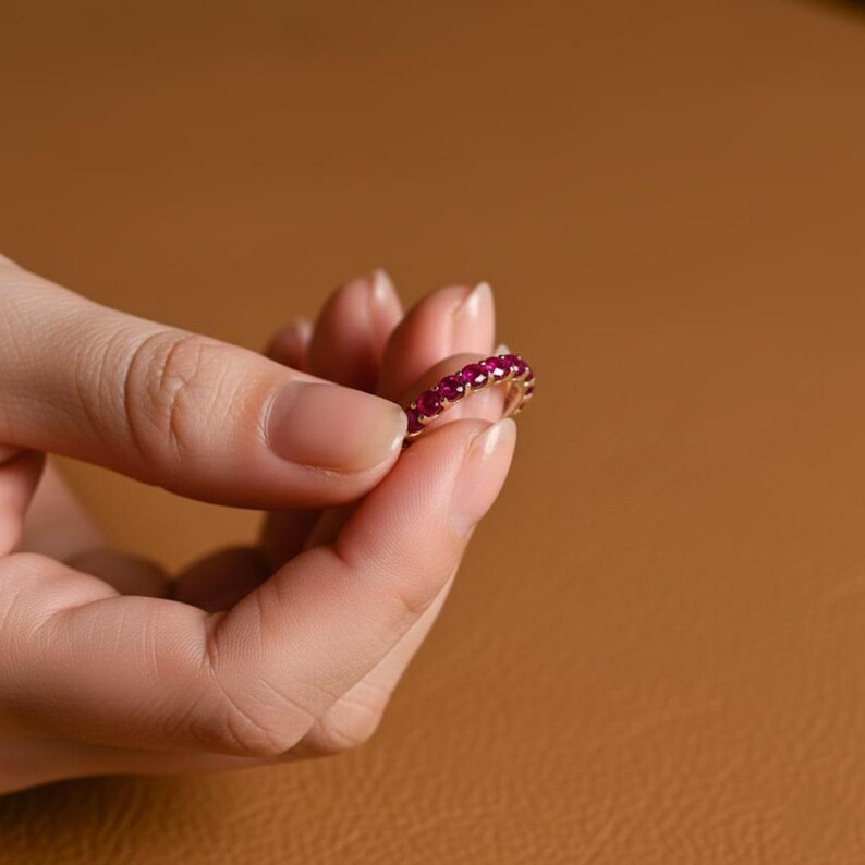 May include: A gold ring with a band of vibrant red gemstones. The ring is held between fingers against a warm brown background, showcasing the ring's design and color. The gemstones are evenly spaced around the ring.