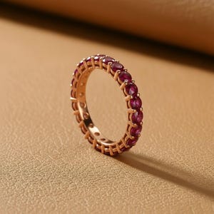 May include: A rose gold eternity ring featuring a continuous row of deep red gemstones. The ring's band is a warm, metallic color, and the gemstones are evenly spaced around the entire circumference, creating a sparkling, elegant look. The ring is set against a neutral background.