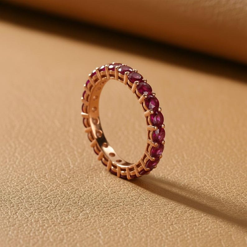 May include: A rose gold eternity ring featuring a continuous row of deep red gemstones. The ring's band is a warm, metallic color, and the gemstones are evenly spaced around the entire circumference, creating a sparkling, elegant look. The ring is set against a neutral background.