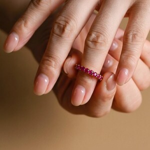 May include: A gold ring set with numerous small, vibrant red gemstones. The ring is worn on a finger, showcasing the band's continuous row of gems. The ring's design is simple, emphasizing the color and sparkle of the stones.