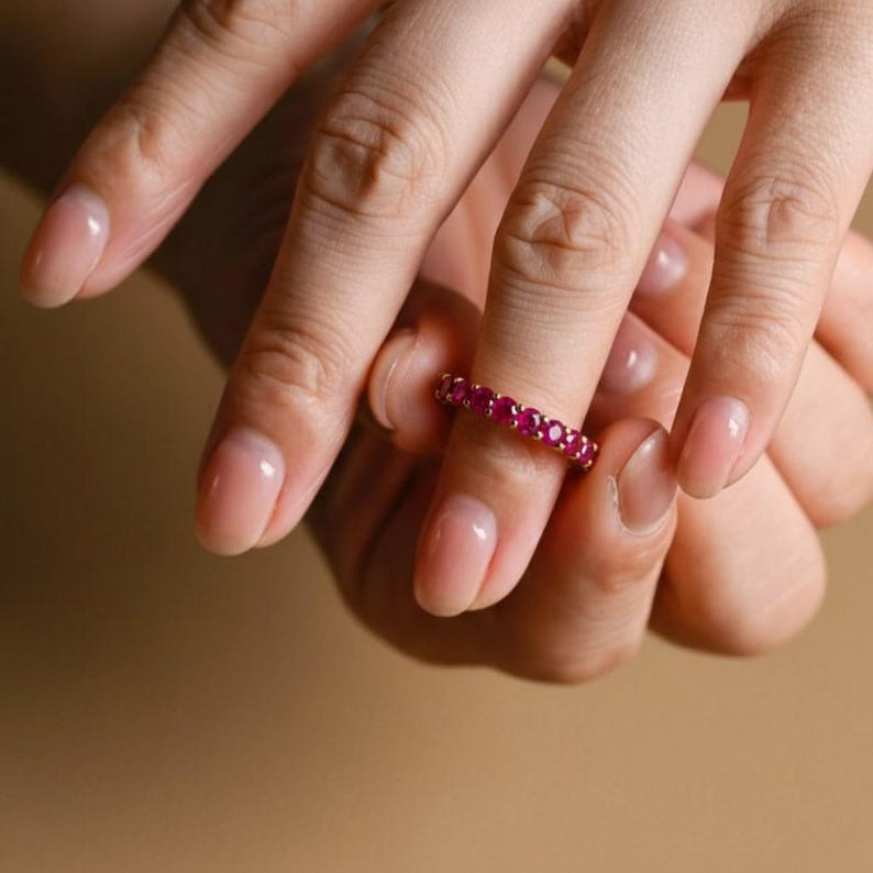 May include: A gold ring set with numerous small, vibrant red gemstones. The ring is worn on a finger, showcasing the band's continuous row of gems. The ring's design is simple, emphasizing the color and sparkle of the stones.
