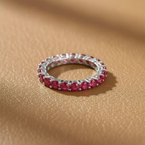 May include: A silver eternity ring with a continuous row of vibrant red gemstones. The ring is set against a warm, neutral background, highlighting the sparkle of the stones. This piece of jewelry is a classic and elegant accessory.