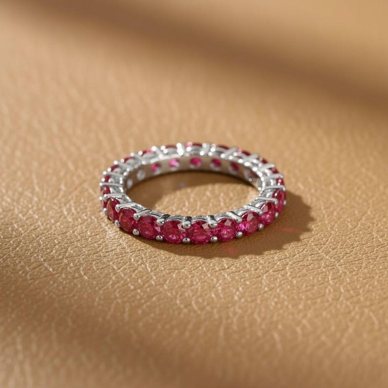 May include: A silver eternity ring with a continuous row of vibrant red gemstones. The ring is set against a warm, neutral background, highlighting the sparkle of the stones. This piece of jewelry is a classic and elegant accessory.