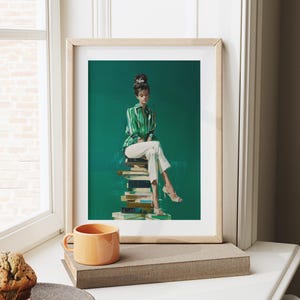 May include: A framed art print featuring a woman in a green and white striped shirt and white pants, seated atop a stack of books against a green backdrop. The print is in a light wood frame. An orange mug sits on a nearby surface.