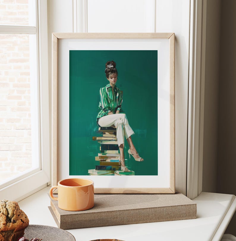 May include: A framed art print featuring a woman in a green and white striped shirt and white pants, seated atop a stack of books against a green backdrop. The print is in a light wood frame. An orange mug sits on a nearby surface.