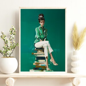 May include: A framed art print depicts a woman in a green and white striped shirt and white pants, seated on a stack of books. The artwork has a green background and the woman is wearing high heels. The print is displayed on a white shelf.