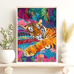 K&ouml;nnte beinhalten: Gerahmter Kunstdruck eines schlafenden Tigers. Der Tiger mit orangefarbenen und schwarzen Streifen liegt auf einer bunten, gemusterten Oberfl&auml;che. Der Hintergrund zeigt &uuml;ppiges gr&uuml;nes Laub.