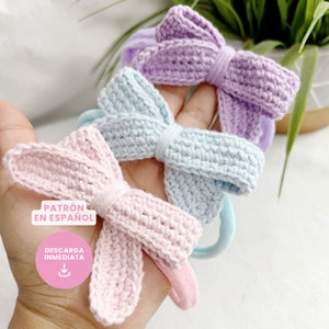 May include: Three crocheted hair bows in pink, light blue and purple. The bows are tied with a knot in the centre and are attached to elastic hair ties. The text "PATRÓN EN ESPAÑOL" and "DESCARGA INMEDIATA" is visible in the image.