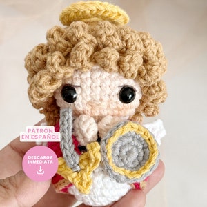 Puede incluir: Un ángel amigurumi tejido a crochet con halo, cabello rubio y un instrumento musical gris y amarillo. El ángel lleva un atuendo rojo y blanco. El texto "PATRÓN EN ESPAÑOL" y "DESCARGA INMEDIATA" es visible en la imagen.
