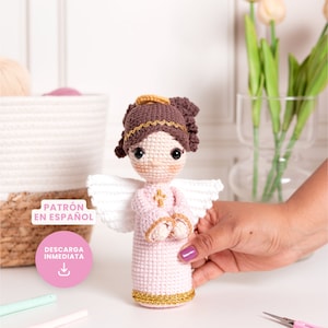 May include: A crocheted angel doll with brown hair, a pink dress, white wings, and a gold halo. The doll is holding its hands together in prayer. The doll is standing on a white surface. The text "PATRÓN EN ESPAÑOL" and "DESCARGA INMEDIATA" is visible in the image.