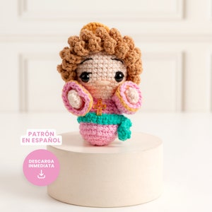 Puede incluir: Una figura de ángel de crochet con cabello castaño, ropa rosa y amarilla y una faja turquesa. El ángel sostiene una pequeña cruz dorada. La figura está sentada sobre un pedestal blanco. El texto "PATRÓN EN ESPAÑOL" y "DESCARGA INMEDIATA" es visible en la imagen.