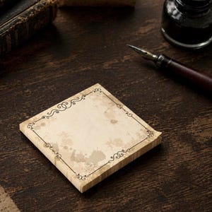 Può includere: Un blocco quadrato di fogli di carta color crema con un motivo di macchie d'inchiostro marrone in stile vintage e un bordo decorativo in filigrana nera. La carta ha un aspetto strutturato ed è posizionata vicino a un pennino in legno scuro e un calamaio.