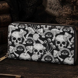 May include: Black and white zippered wallet featuring a repeating pattern of skulls, mushrooms, and eyes. The wallet has a gold-colored zipper and a rectangular shape. The design is gothic and whimsical.