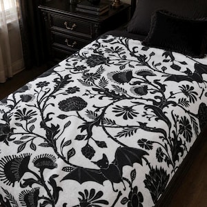 Könnte beinhalten: Eine weiße Wohndecke mit einem sich wiederholenden schwarzen Muster aus stilisierten Blumen, blättrigen Ranken und kleinen Fledermäusen mit ausgebreiteten Flügeln. Der Stoff hat eine weiche Textur und bedeckt ein Bett mit schwarzem Kissen.