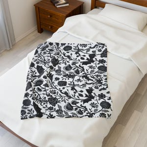 Puede incluir: Una manta estampada en blanco y negro con un diseño floral y de murciélagos. La manta está colocada sobre una cama blanca, con una mesita de noche de madera en el fondo. La manta rectangular parece estar hecha de material suave.