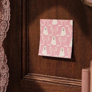 May include: A square sticky note featuring a repeating pattern of white ghosts and light pink ribbons on a solid pink background. Each ghost has two black eyes and a small black heart-shaped mouth, while the ribbons are depicted in a simple line art style.