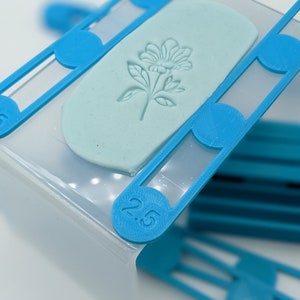 Könnte beinhalten: Ein blaues Plastikwerkzeug mit einem eingeprägten Blumendesign auf einer hellblauen Tonfläche. Das Werkzeug hat die Nummer "2,5" aufgedruckt.