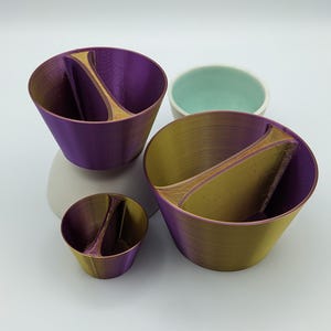 Pode incluir: Três tigelas de tamanhos diferentes, impressas em 3D, com um design único dividido. As tigelas são roxas, douradas e um tom roxo mais claro. As tigelas são feitas de um plástico liso e brilhante.