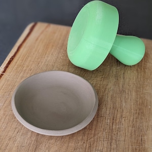 Puede incluir: Un molde de cer&aacute;mica verde claro para hacer cuencos peque&ntilde;os, junto a un cuenco de arcilla gris sobre una superficie de madera.