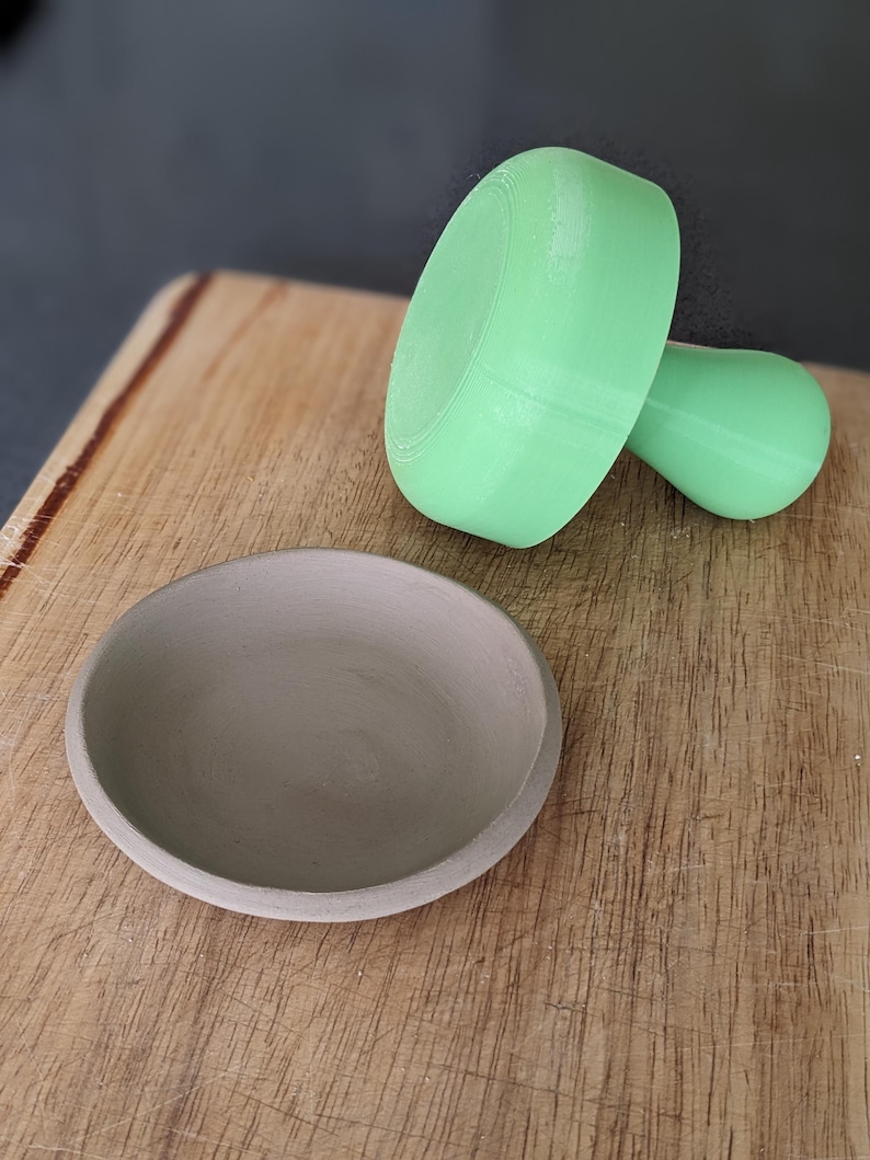Puede incluir: Un molde de cer&aacute;mica verde claro para hacer cuencos peque&ntilde;os, junto a un cuenco de arcilla gris sobre una superficie de madera.