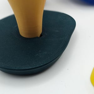 Peut inclure: Une base ovale de couleur teal avec un objet conique de couleur moutarde ins&eacute;r&eacute;. La base a une surface textur&eacute;e. Le c&ocirc;ne est centr&eacute; et a une finition lisse. Le fond est blanc.
