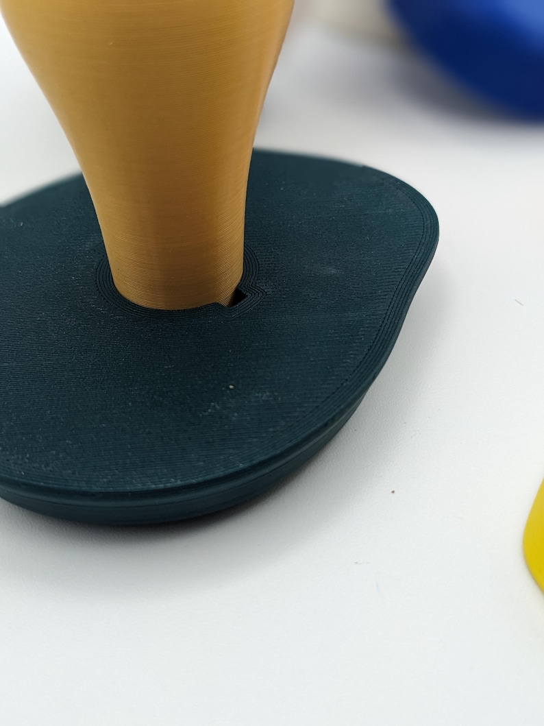 Peut inclure: Une base ovale de couleur teal avec un objet conique de couleur moutarde ins&eacute;r&eacute;. La base a une surface textur&eacute;e. Le c&ocirc;ne est centr&eacute; et a une finition lisse. Le fond est blanc.