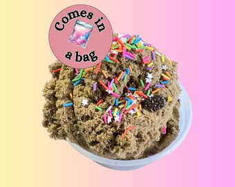 May include: A textured brown substance resembling cookie dough in a white bowl, topped with colourful sprinkles and small white star-shaped pieces. A circular pink sticker with the text "Comes in a bag" and an illustration of a bag is attached.