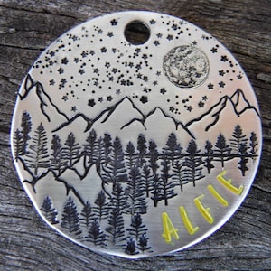 Puede incluir: Una placa redonda plateada para mascotas con una escena de montaña y bosque bajo un cielo estrellado y una luna llena. El nombre "ALFIE" está escrito en letras amarillas en la parte inferior. La placa tiene un agujero en la parte superior para sujetarla.