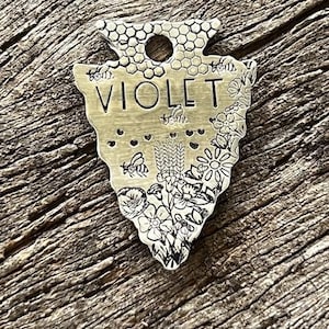 Può includere: Un'etichetta per animali domestici color ottone a forma di punta di freccia, incisa con il nome "VIOLET". L'etichetta presenta disegni floreali e di api, con un motivo a nido d'ape nella parte superiore. L'etichetta è su una superficie di legno stagionato.