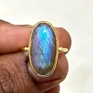 May include: A gold-colored ring featuring an oval-shaped labradorite stone. The stone displays a mix of blue, purple, and gray hues. The ring has a simple, elegant design, with the stone set in a gold bezel.