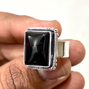 Op de afbeelding: Een vierkante zwarte onyx edelsteen ring met een zilveren band. De onyx is gezet in een rechthoekige vorm met een decoratieve rand. De ring wordt tegen een witte achtergrond gehouden.