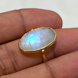 Peut inclure: Bague en or ornée d'une pierre de lune ovale et irisée, affichant des reflets bleus et blancs. La monture et le sertissage sont dorés, créant un design classique et élégant. Bijou.
