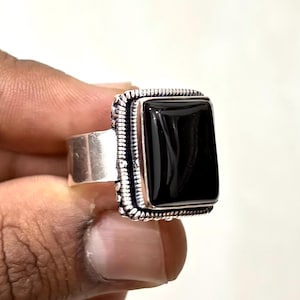 May include: A silver ring with a large, rectangular black stone. The stone is set in a decorative bezel with a textured border. The ring is held in a hand against a white background. The ring is a piece of jewelry.