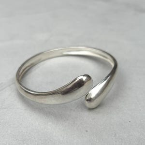May include: A silver-colored ring with a minimalist design. The ring features a slender band that curves into two teardrop-shaped ends, creating an open, adjustable style. The ring is made of a shiny, reflective metal.