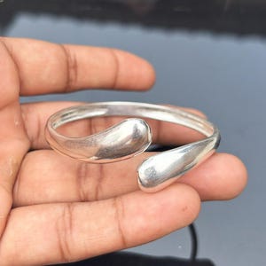 May include: A silver-colored cuff bracelet with an open design. The bracelet features two teardrop-shaped terminals. The jewelry is held in a hand, showcasing the polished metal and simple, elegant style. The bracelet is a piece of fashion jewelry.