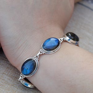 Könnte beinhalten: Ein silbernes Armband mit drei ovalen Labradoritsteinen. Die Steine zeigen schillernde blaue und grüne Farbtöne. Das Armband ist durch silberne Glieder verbunden und wird am Handgelenk getragen.