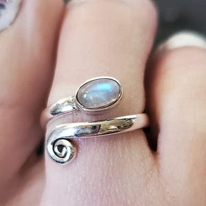 Puede incluir: Un anillo de plata con una piedra de luna engastada en un diseño de espiral. El anillo está en un dedo.