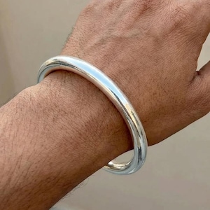 Può includere: Un braccialetto a cerchio argentato lucido. Il braccialetto è una fascia solida e arrotondata. La superficie liscia e riflettente del braccialetto cattura la luce. Il braccialetto è indossato al polso.