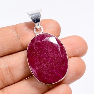 Puede incluir: Un colgante de rubí con una gema ovalada pulida. La piedra de color rojo intenso está engastada en un marco de metal plateado con un pequeño lazo para una cadena. El colgante se sostiene en una mano sobre un fondo blanco.