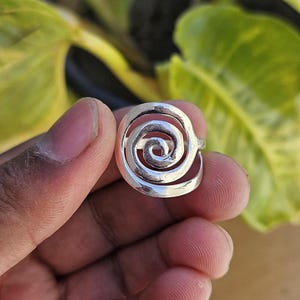 Peut inclure: Une bague argentée avec un motif en spirale. Le point central de la bague est un motif tourbillonnant, créant une sensation de mouvement. La bague est tenue dans une main, avec un arrière-plan vert flou.